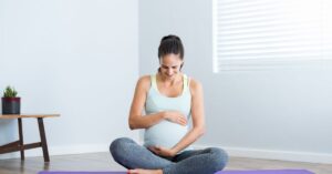 Pregnant woman sitting cross-legged, smiling and holding her baby bump while practicing affirmations