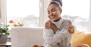Smiling woman hugging herself while sitting on a sofa, symbolizing self-love and inner peace.